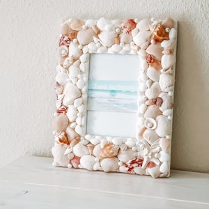 May include: White picture frame decorated with seashells and pearls. The frame is rectangular and has a white background. The text "Ombre Sea Designs" is visible in the frame.