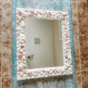 May include: A white mirror with a decorative frame made of seashells. The frame is made of a variety of seashells, including white, pink, and brown shells. The mirror is hanging on a wall with a blue and white patterned rug.