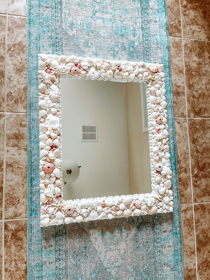 May include: A white mirror with a decorative frame made of seashells. The frame is made of a variety of seashells, including white, pink, and brown shells. The mirror is hanging on a wall with a blue and white patterned rug.