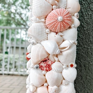 May include: A white mirror frame decorated with seashells and pearls. The frame is made of various types of seashells, including scallop shells, conch shells, and sand dollars. The shells are arranged in a random pattern and are held together with glue. The mirror is surrounded by a border of white pearls.