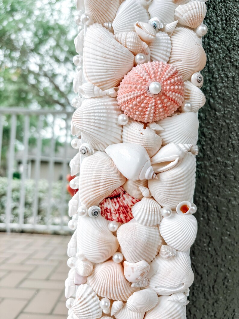 May include: A white mirror frame decorated with seashells and pearls. The frame is made of various types of seashells, including scallop shells, conch shells, and sand dollars. The shells are arranged in a random pattern and are held together with glue. The mirror is surrounded by a border of white pearls.
