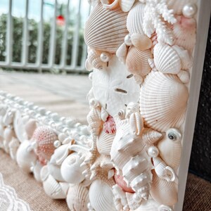 May include: A white mirror frame decorated with seashells and pearls. The frame is made of wood and has a white finish. The seashells are arranged in a random pattern and are of various sizes and shapes. The pearls are scattered throughout the frame and add a touch of elegance.
