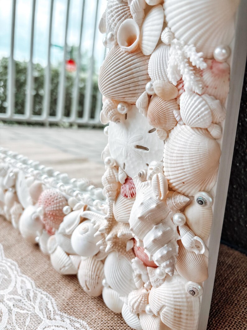May include: A white mirror frame decorated with seashells and pearls. The frame is made of wood and has a white finish. The seashells are arranged in a random pattern and are of various sizes and shapes. The pearls are scattered throughout the frame and add a touch of elegance.
