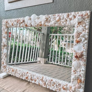 May include: Rectangular mirror with a white frame lavishly decorated with various seashells in shades of white, beige, and brown. The mirror is propped up on a beige surface, reflecting a balcony with white railings and greenery.