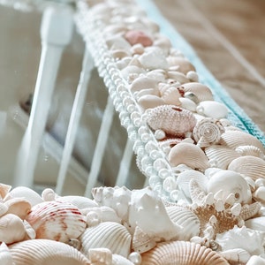 May include: A white mirror with a decorative border made of seashells and pearls. The seashells are in various shapes and sizes, and some are painted in shades of pink and white.