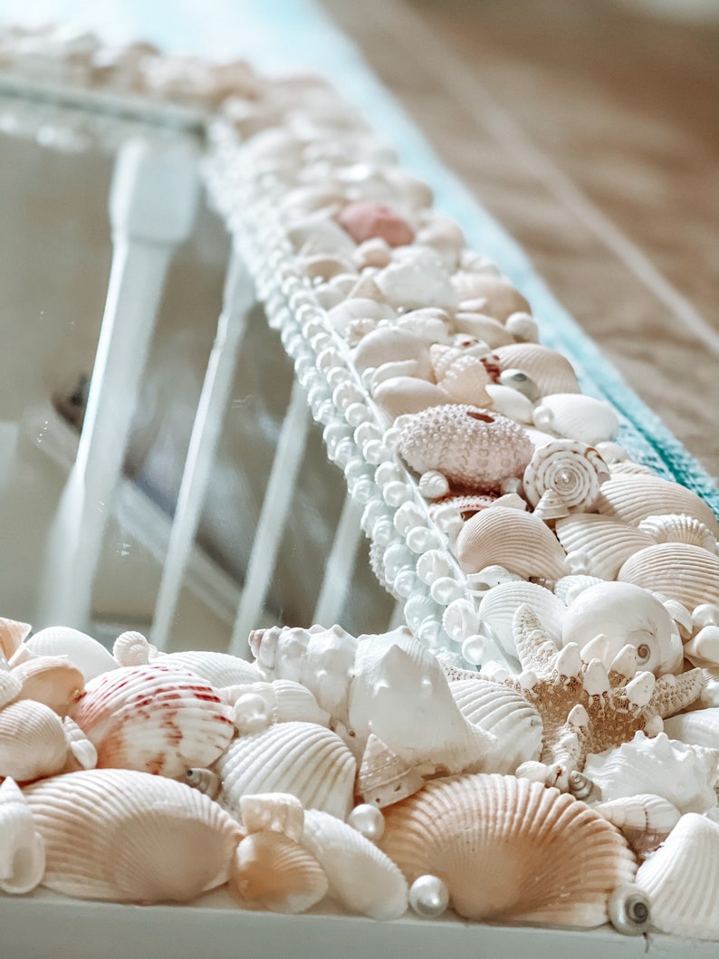 May include: A white mirror with a decorative border made of seashells and pearls. The seashells are in various shapes and sizes, and some are painted in shades of pink and white.