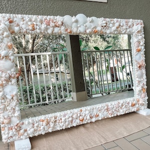 May include: A large rectangular mirror with a frame made entirely of seashells. The shells are mostly white with some pink and peach accents. The mirror is reflecting a view of a patio with a metal fence and trees.