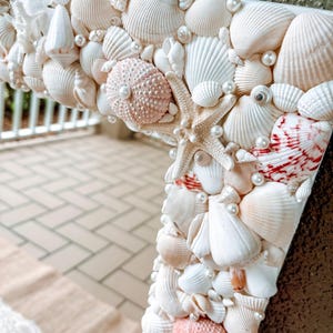May include: A white mirror frame decorated with seashells and pearls. The frame is made of various sizes and shapes of seashells, including scallop shells, conch shells, and sand dollars. The frame is accented with small white pearls.