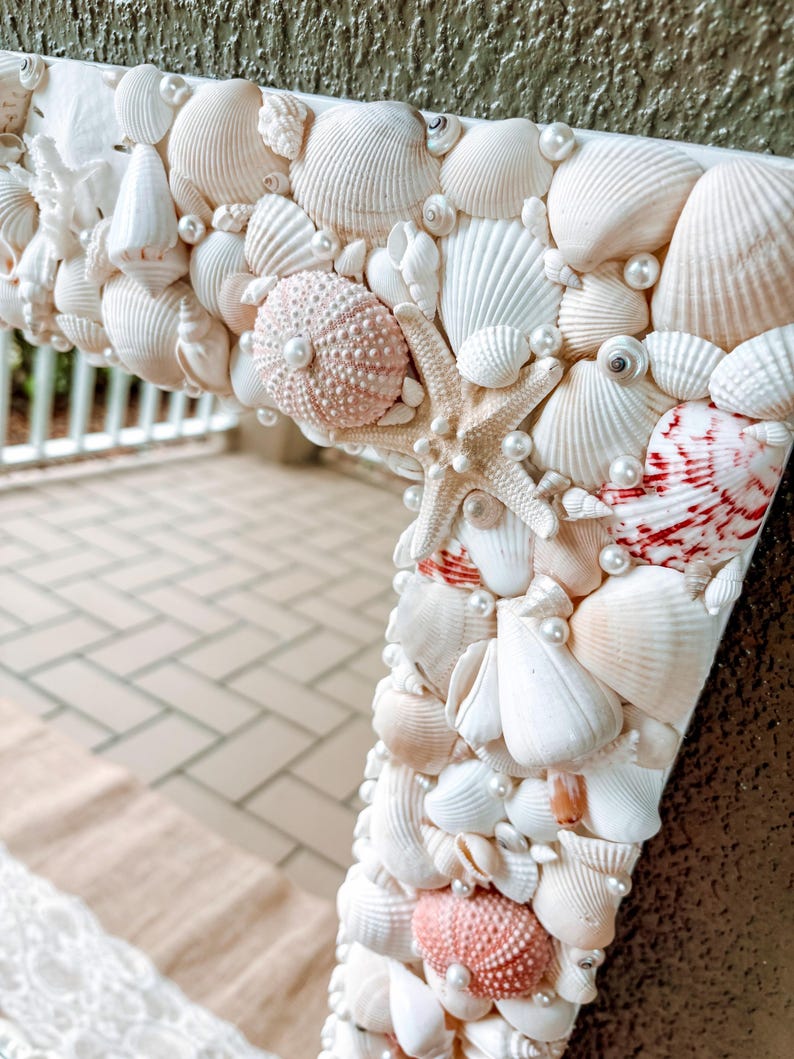 May include: A white mirror frame decorated with seashells and pearls. The frame is made of various sizes and shapes of seashells, including scallop shells, conch shells, and sand dollars. The frame is accented with small white pearls.