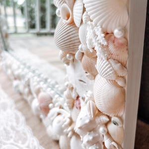 May include: A white mirror frame decorated with seashells and pearls. The frame is made of wood and has a white finish. The seashells are arranged in a random pattern and are of various sizes and shapes. The pearls are scattered throughout the frame and add a touch of elegance.