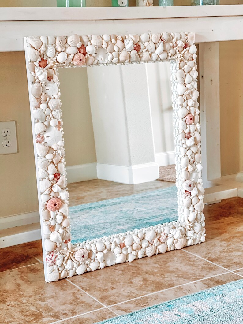 May include: A white mirror with a frame made of seashells. The frame is decorated with pink and white seashells.