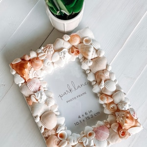 May include: A white photo frame decorated with seashells. The frame is 4 x 6 inches (10.1 x 15.2 cm) and has the text "parklane PHOTO FRAME" printed on it.