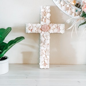 May include: A white wooden cross decorated with seashells. The center of the cross features a large pink seashell.