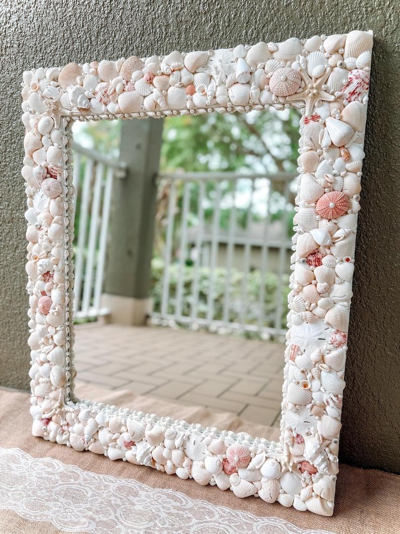May include: A white mirror with a decorative frame made of seashells. The frame is made of a variety of seashells, including white, pink, and brown shells. The mirror is reflecting a blurry image of a patio.