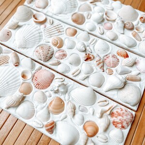 May include: Three white rectangular panels with a variety of seashells glued to the surface. The shells are in various sizes and colors, including white, brown, and pink.