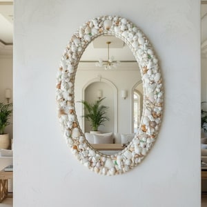 May include: Oval mirror with a frame decorated with various seashells in shades of white, beige, and brown. The mirror is mounted on a white wall, reflecting a bright, airy interior with neutral tones and natural light.