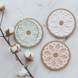 Peut inclure: Trois dessous de verre décoratifs avec des motifs floraux. Un est bleu clair, un blanc et un rose. Chacun a une base en liège et un motif floral blanc en relief. Une brindille de coton ajoute une touche naturelle.
