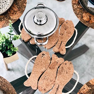 May include: A stainless steel pot with a lid sits atop cork fish-shaped trivets. The trivets are arranged on a glass-topped table, alongside cork placemats and a small potted plant. The fish trivets have white rope handles.