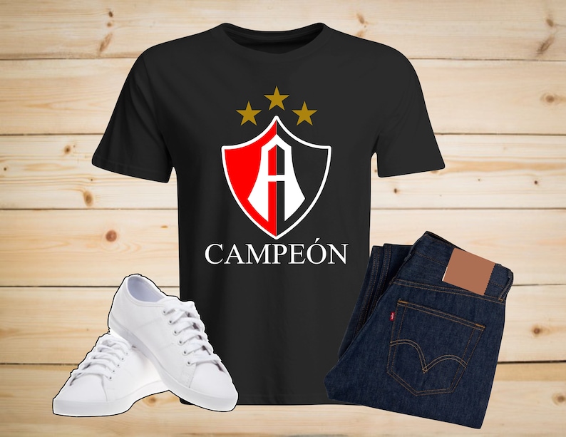 Puede incluir: Camiseta negra con un escudo rojo y blanco que presenta una letra "A" y cuatro estrellas doradas. El texto "CAMPE&Oacute;N" est&aacute; impreso debajo del escudo.