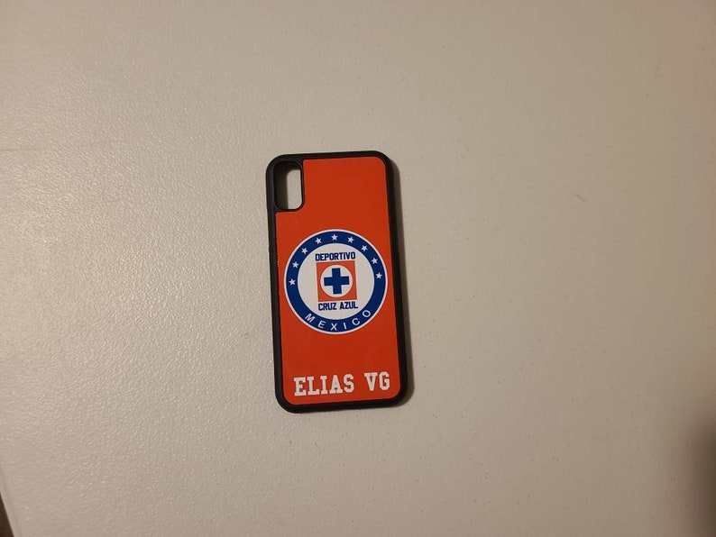 May include: An orange phone case with a black border. The case features the Deportivo Cruz Azul logo in a blue and white circular design. The name "ELIAS VG" is printed in white at the bottom of the case.