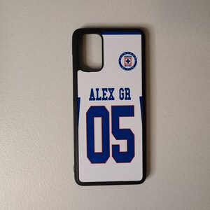 May include: A white phone case with a blue and black design. The case features the text "ALEX GR" above the number "05" in blue. A team logo is in the upper right corner. The case has a black border.