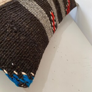 K&ouml;nnte beinhalten: Nahaufnahme eines schwarzen, grauen, wei&szlig;en und roten gestreiften Kissens mit blau-wei&szlig;em Besatz. Das Kissen ist aus gewebtem Stoff und hat einen rustikalen, handgefertigten Look.
