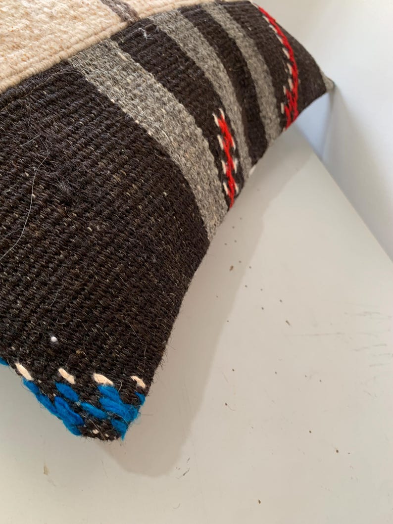 K&ouml;nnte beinhalten: Nahaufnahme eines schwarzen, grauen, wei&szlig;en und roten gestreiften Kissens mit blau-wei&szlig;em Besatz. Das Kissen ist aus gewebtem Stoff und hat einen rustikalen, handgefertigten Look.