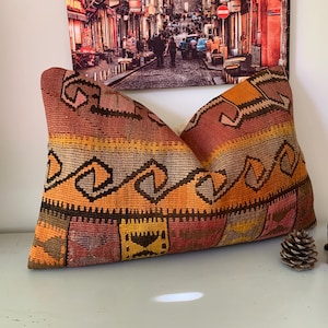 May include: A rectangular, patterned pillow with a warm color palette of orange, yellow, and brown. The pillow features geometric designs and is placed on a white surface. A wooden elephant figurine and a pine cone are next to the pillow.