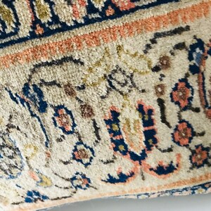 May include: Close-up of a cream-colored pillow with a blue, pink, and gold floral pattern. The pillow has a fringe border.
