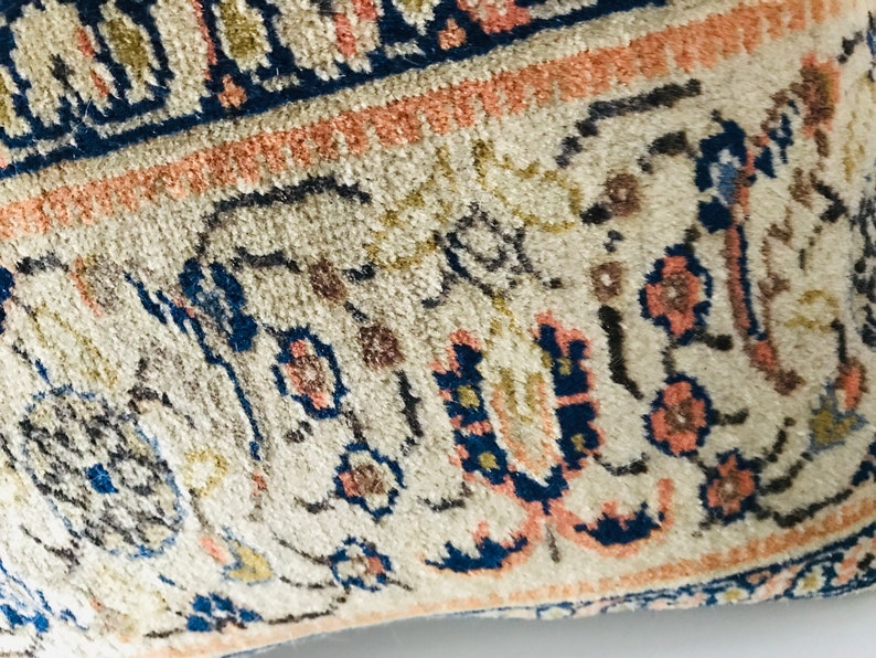 May include: Close-up of a cream-colored pillow with a blue, pink, and gold floral pattern. The pillow has a fringe border.