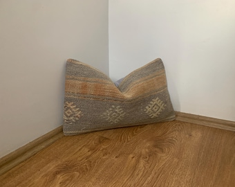 May include: Rectangular cushion with a woven texture. The cushion features a colour palette of grey, beige, and orange hues. It has a geometric pattern in white and is placed in a corner against a white wall and wooden floor.