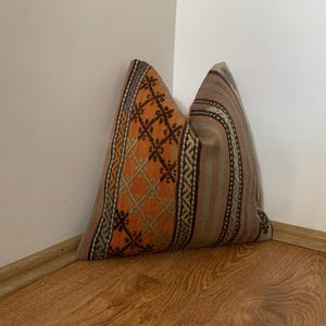 May include: A decorative pillow with a geometric pattern in orange, brown, and beige. The pillow has a striped design on one side and is placed in a corner against a white wall and wooden floor.