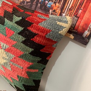 May include: A colorful throw pillow with a geometric pattern in shades of red, green, black, and white. The pillow is made of a woven fabric and has a fringed edge.