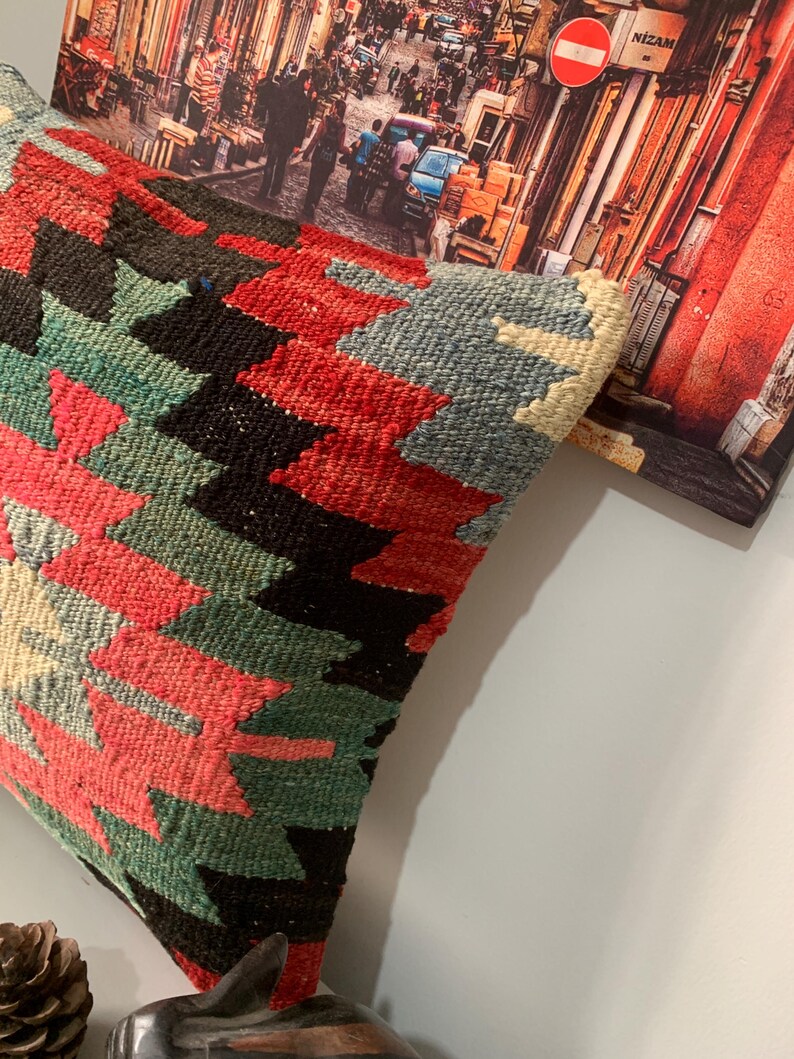May include: A colorful throw pillow with a geometric pattern in shades of red, green, black, and white. The pillow is made of a woven fabric and has a fringed edge.