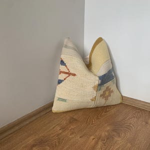 May include: A decorative pillow with a patchwork design. The pillow features a cream-colored base with geometric patterns in blue, orange, and green. The top corner is yellow, and the pillow is resting on a wooden floor.