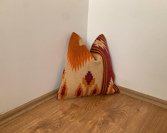 May include: A square throw pillow featuring a woven textile pattern in shades of cream, burnt orange, and deep maroon. The design includes geometric shapes and stylized floral motifs arranged in vertical bands across the front surface.