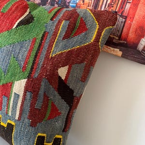 May include: A colorful, geometric patterned throw pillow with a blue, green, red, black, and yellow design. The pillow is made of woven fabric and has a fringed edge.