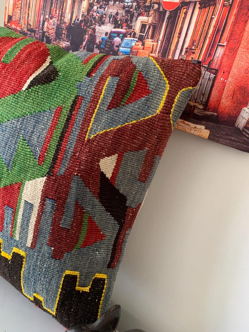 May include: A colorful, geometric patterned throw pillow with a blue, green, red, black, and yellow design. The pillow is made of woven fabric and has a fringed edge.