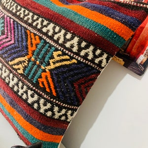 May include: A colorful throw pillow with a geometric pattern in shades of red, orange, blue, green, and white. The pillow is made of woven fabric and has a fringe border.