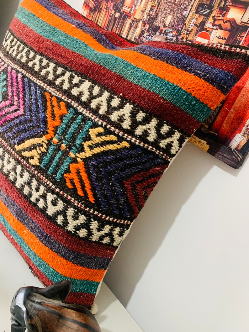 May include: A colorful throw pillow with a geometric pattern in shades of red, orange, blue, green, and white. The pillow is made of woven fabric and has a fringe border.