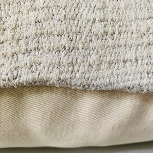 May include: Close-up of a beige throw pillow with a textured fabric. The pillow is made of two layers of fabric, with the top layer being a woven fabric and the bottom layer being a smooth fabric.