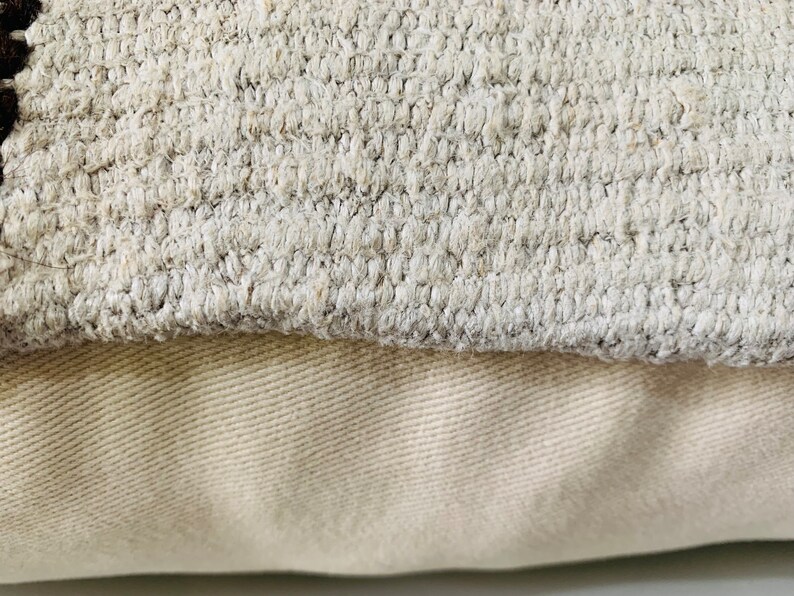 May include: Close-up of a beige throw pillow with a textured fabric. The pillow is made of two layers of fabric, with the top layer being a woven fabric and the bottom layer being a smooth fabric.