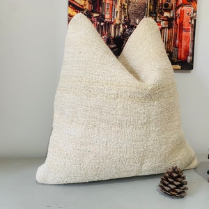 Peut inclure: Oreiller texturé de couleur crème de forme triangulaire. L'oreiller est fait d'un tissu tissé et a un aspect doux et accueillant. Un éléphant en bois décoratif et une pomme de pin sont à côté de l'oreiller.