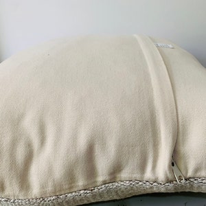 May include: A white pillow with a zipper closure and a beige woven trim around the edges.