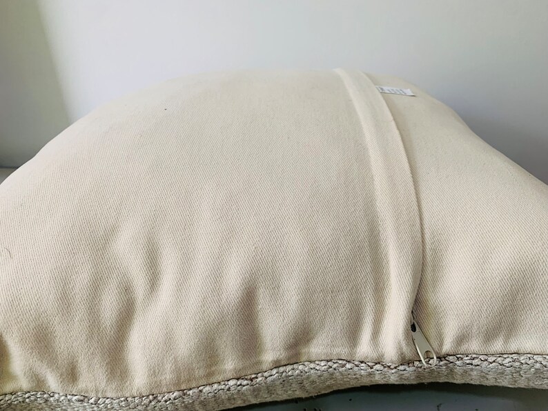 May include: A white pillow with a zipper closure and a beige woven trim around the edges.
