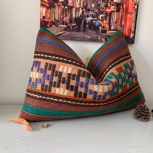 May include: A decorative pillow with a colorful geometric pattern in shades of brown, green, blue, and orange. The pillow is made of woven fabric and has a rectangular shape.