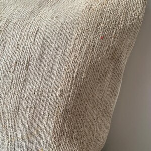 May include: Close-up of a beige woven pillow with a textured surface. The pillow has a neutral color palette and a rustic aesthetic. The pillow is likely used for home decor.
