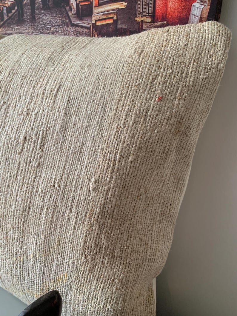 May include: Close-up of a beige woven pillow with a textured surface. The pillow has a neutral color palette and a rustic aesthetic. The pillow is likely used for home decor.