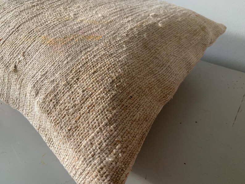 May include: A beige woven pillow with a textured surface. The pillow has a square shape and is made of a natural fiber material. The weave pattern is visible, creating a rustic aesthetic. The pillow is a decorative home accent.
