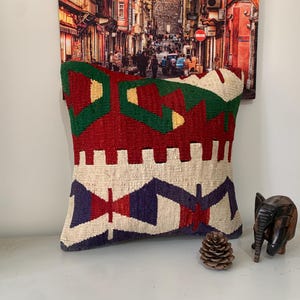 May include: A colorful throw pillow with a geometric pattern in shades of red, green, white, and blue. The pillow is made of woven fabric and has a fringed edge.